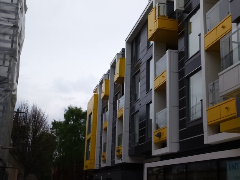 Modern apartment building with dark gray cladding and bright yellow balcony boxes, trees in background, overcast sky