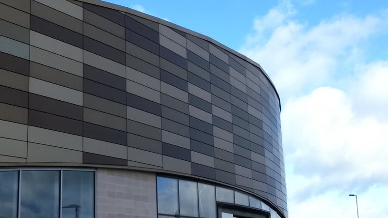 Curved building facade with gray and white checkered panel pattern under a blue sky with clouds