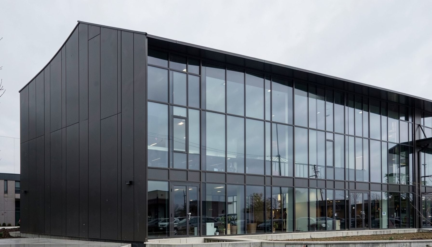 Modern commercial building facade with dark metal cladding panels and large glazed curtain wall, overcast sky