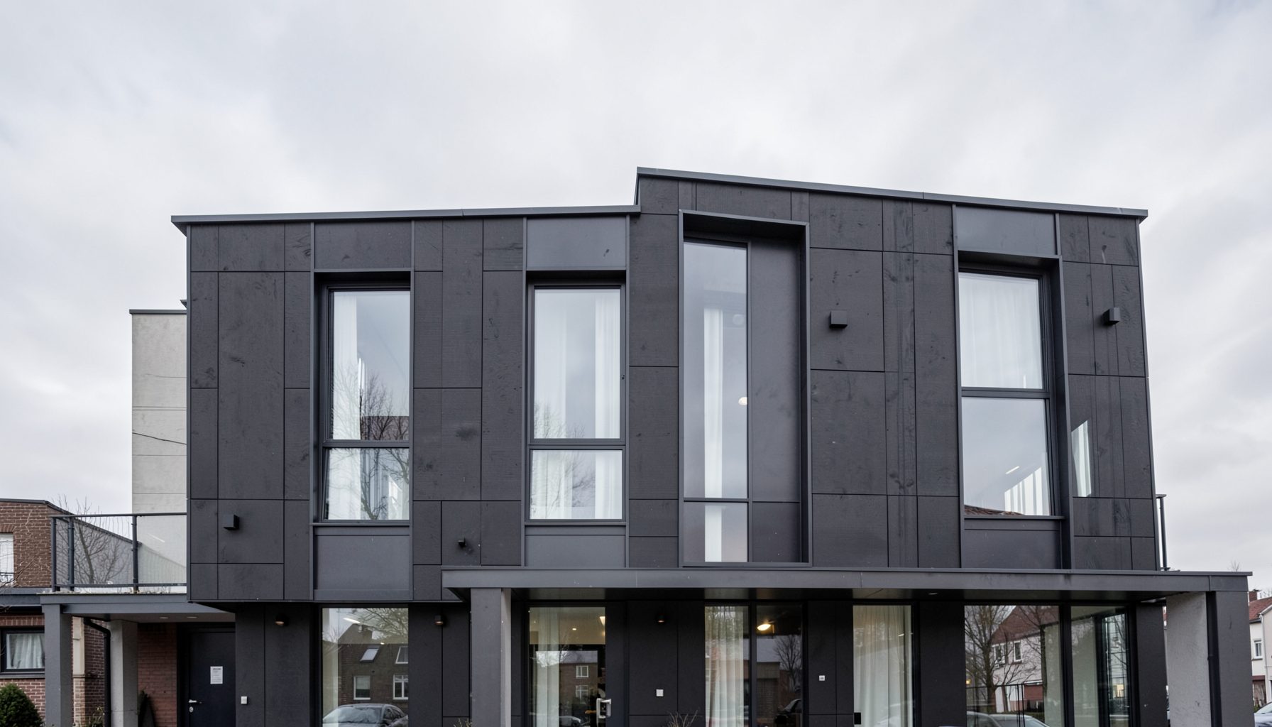 Modern residential building facade with dark cladding panels and large windows, set against an overcast sky