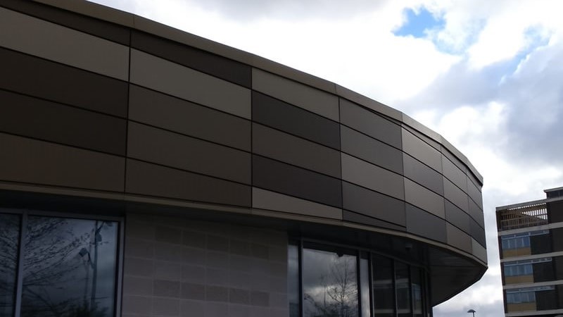 Curved building facade with brown and beige geometric panel pattern under a partly cloudy sky