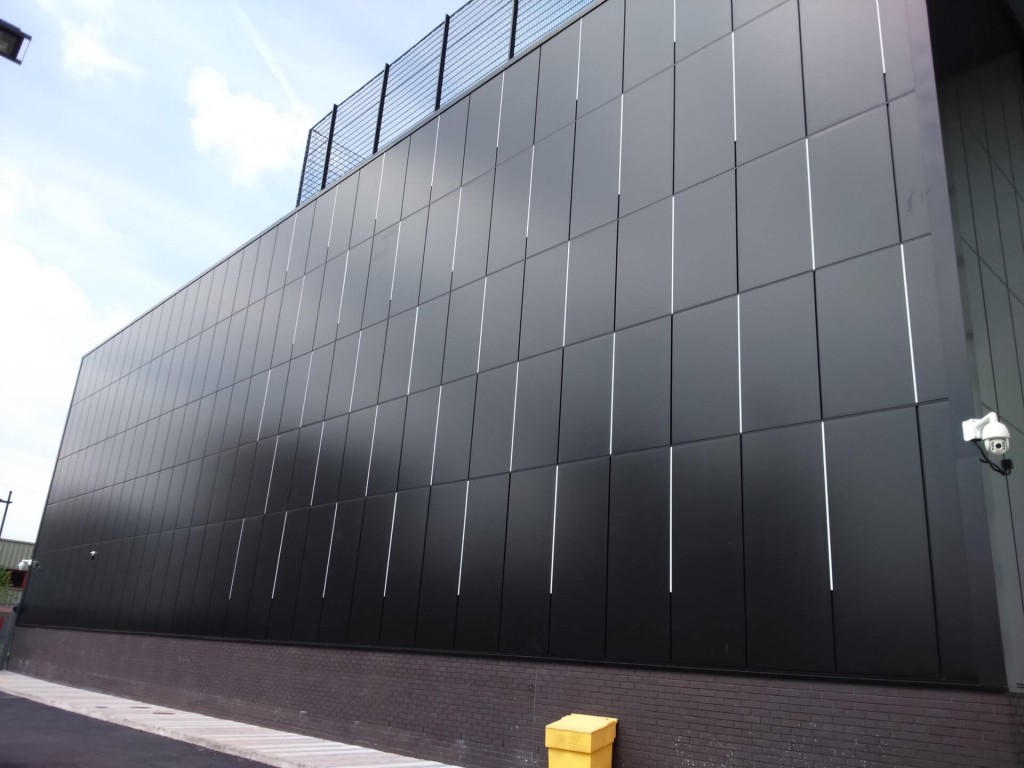 Large dark metal-paneled wall of a building with vertical light-colored seams, security camera mounted, cloudy blue sky