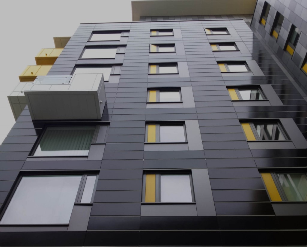Upward view of a modern high-rise apartment building facade with dark gray panels, yellow accent strips, and balconies.