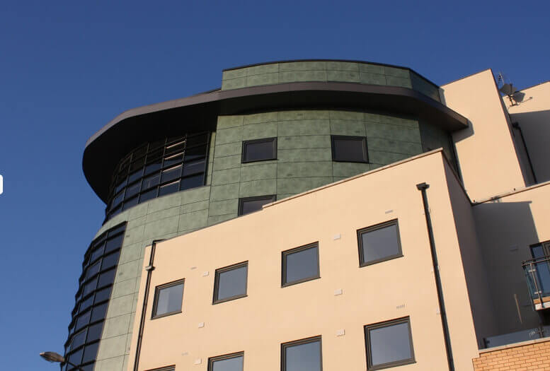 Curved modern building with green paneled upper section and beige lower section, windows, under a clear blue sky
