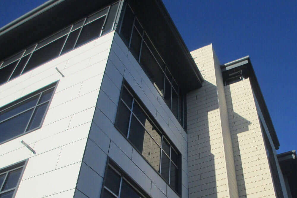 Upward angled view of a modern building corner with cream and dark paneled cladding, large windows, against a deep blue sky