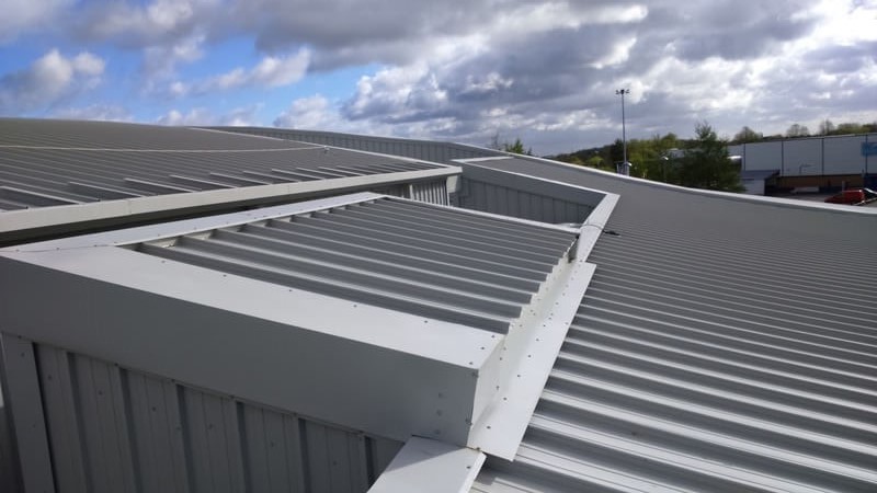 Close-up of grey metal roof panels with ridged sections and roof vents against a partly cloudy sky, industrial and muted mood